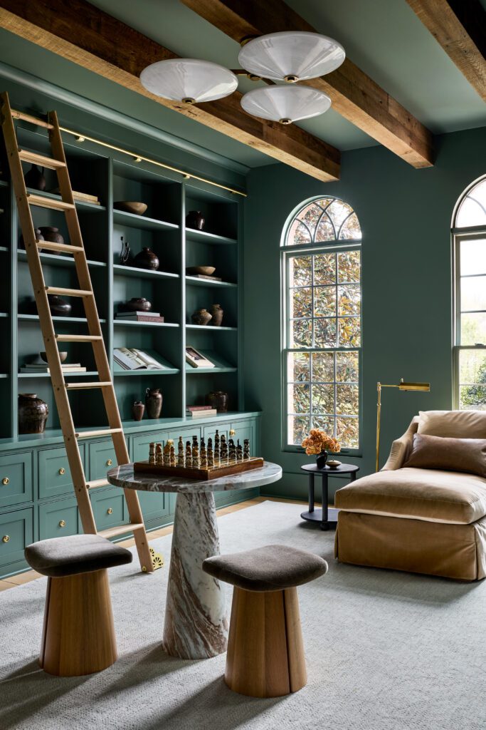 Custom floor-to-ceiling green library cabinetry with rolling ladder, brass hardware, exposed wood beams, and arched windows by Unique Kitchens & Baths™.
