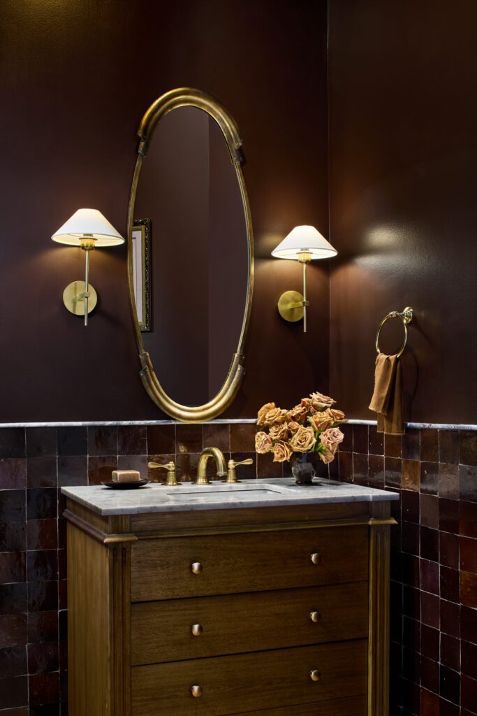 Furniture-style wood bathroom vanity with marble countertop, brass oval mirror, sconces, and deep burgundy tile by Unique Kitchens & Baths™.