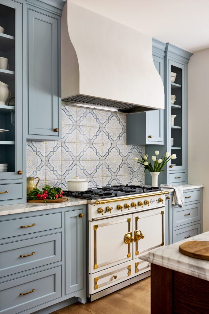 Custom blue kitchen cabinetry with brass hardware, patterned tile backsplash, statement range, and sculptural plaster hood by Unique Kitchens & Baths™.