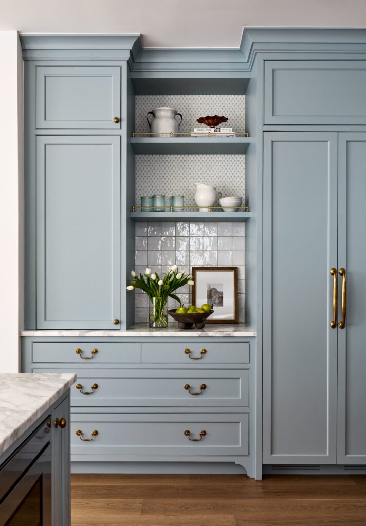 Custom blue-gray kitchen cabinetry with brass hardware, open shelving, tiled backsplash, and marble countertops by Unique Kitchens & Baths™.