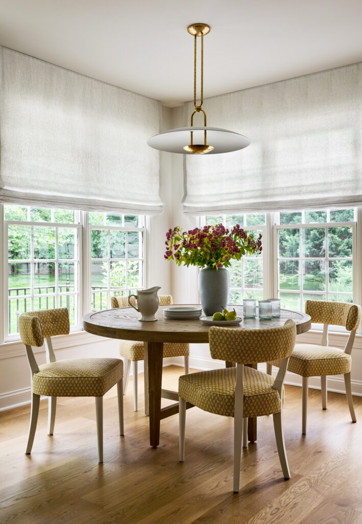 Bright kitchen dining nook with round wood table, upholstered chairs, linen window treatments, and brass pendant lighting.
