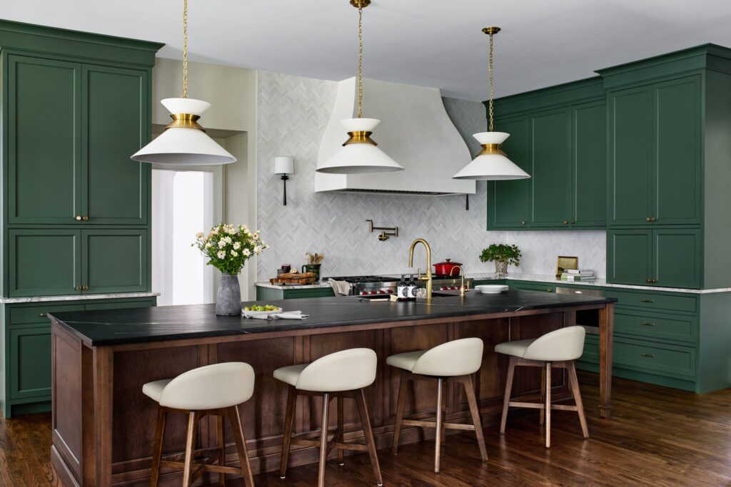Custom green kitchen cabinetry with large dark wood island, black countertops, brass fixtures, statement pendant lights, and sculptural range hood by Unique Kitchens & Baths™.