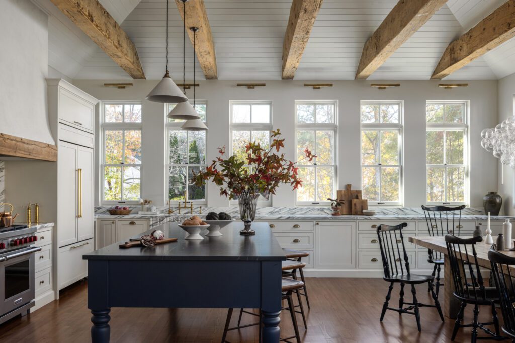 Timeless kitchen with custom white cabinetry, navy island, marble countertops, exposed wood beams, and abundant natural light by Unique Kitchens & Baths™.